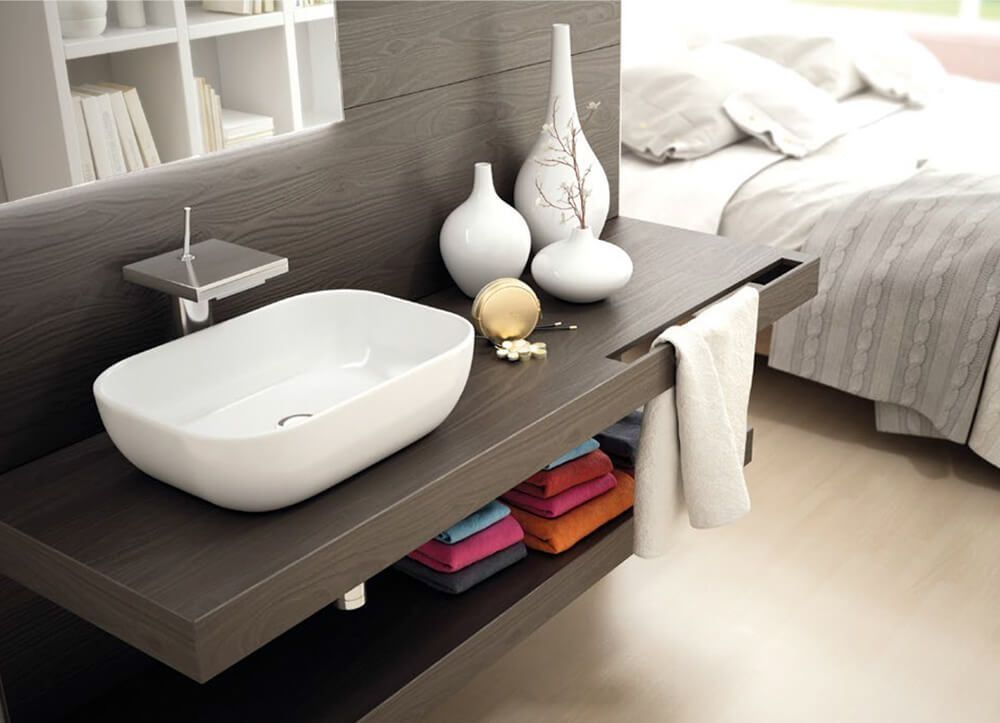 Tocador de baño moderno con lavabo blanco, estante y jarrones decorativos frente a una cama.
