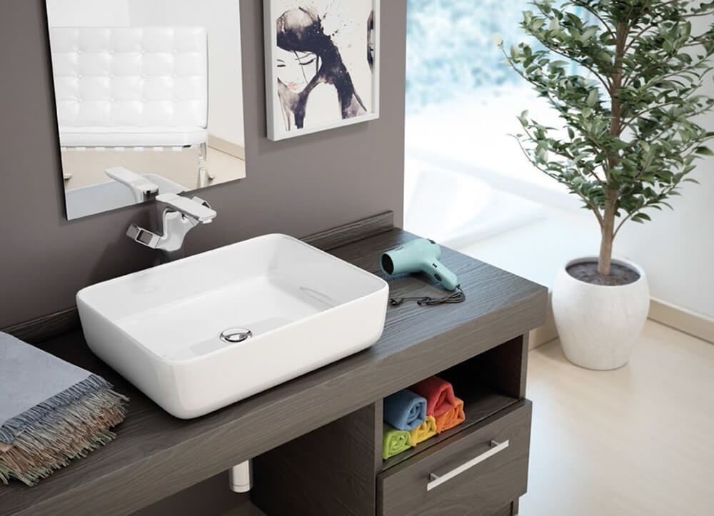 Baño con lavabo rectangular blanco, espejo, arte, secador de pelo y planta sobre encimera gris.