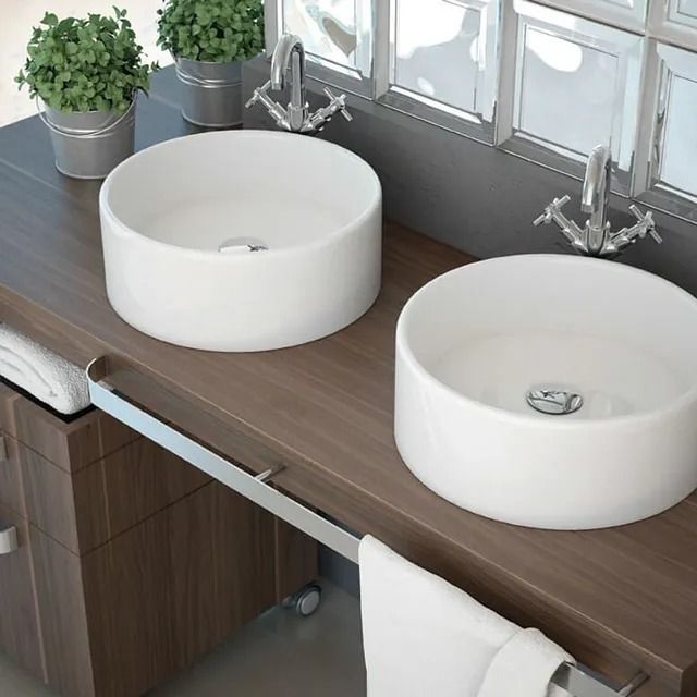 Dos lavabos circulares blancos sobre un tocador de madera en un baño con accesorios cromados y pequeñas plantas en macetas.
