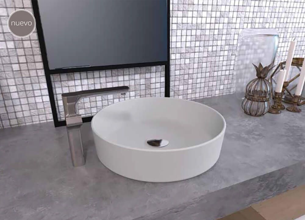 Lavabo de baño circular de color blanco sobre encimera gris con grifo y fondo de mosaico.