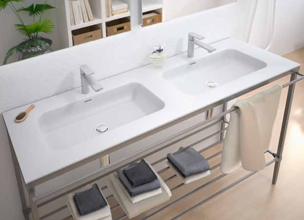 Lavabo doble blanco sobre estructura metálica, toallas y artículos de aseo.