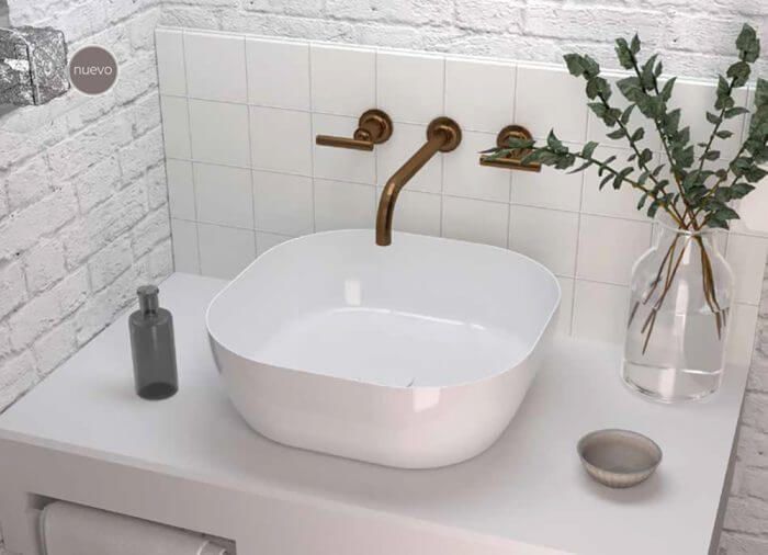 Lavabo de baño blanco con grifo de latón, jabón y vegetación sobre encimera blanca sobre un fondo estilo ladrillo.