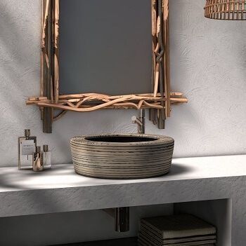 Baño con espejo con marco de madera, lavabo redondo texturizado sobre encimera blanca, con artículos de aseo y pantalla de lámpara tejida.