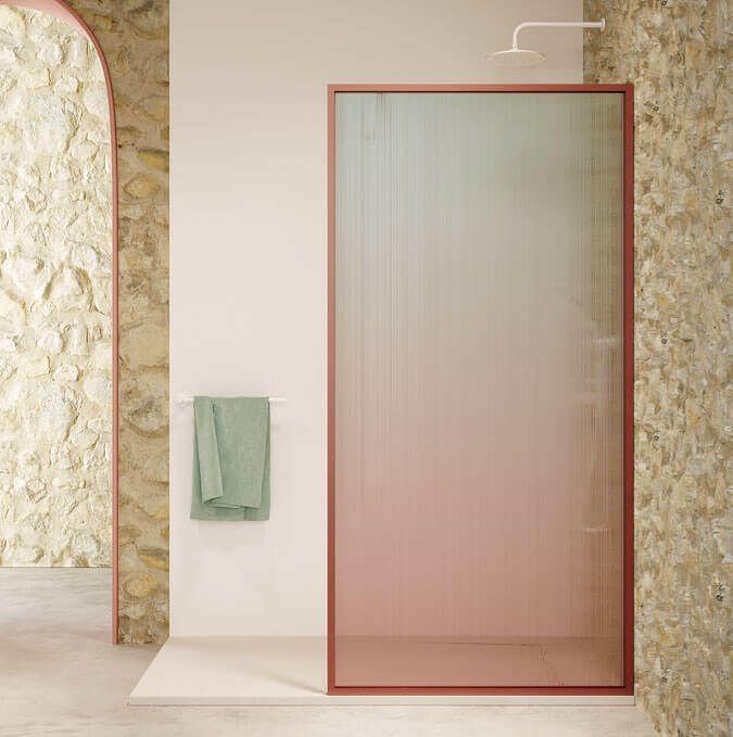 Ducha con puerta de cristal con marco rosa, efecto degradado, en un baño con paredes de piedra y toalla verde.