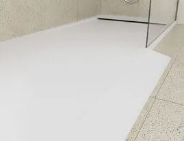Base de ducha de color blanco, parte de una ducha con mampara de cristal, junto a un suelo y una pared de baldosas de color claro.