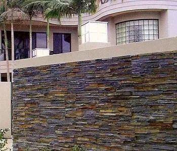 Muro revestido de piedra frente a una casa moderna con grandes ventanales y palmeras.