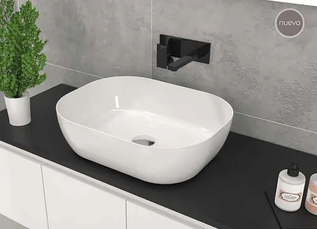 Lavabo ovalado blanco sobre encimera negra, grifo negro, pequeña planta, dispensador de jabón y pared gris.