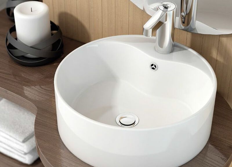 Lavabo de baño redondo blanco con grifo plateado, sobre encimera de madera, vela en portavelas.
