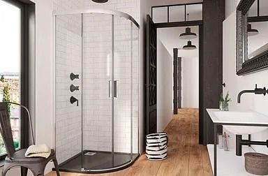 Baño moderno con ducha con marco negro, tocador flotante de madera, espejo circular, paredes grises y un inodoro blanco.