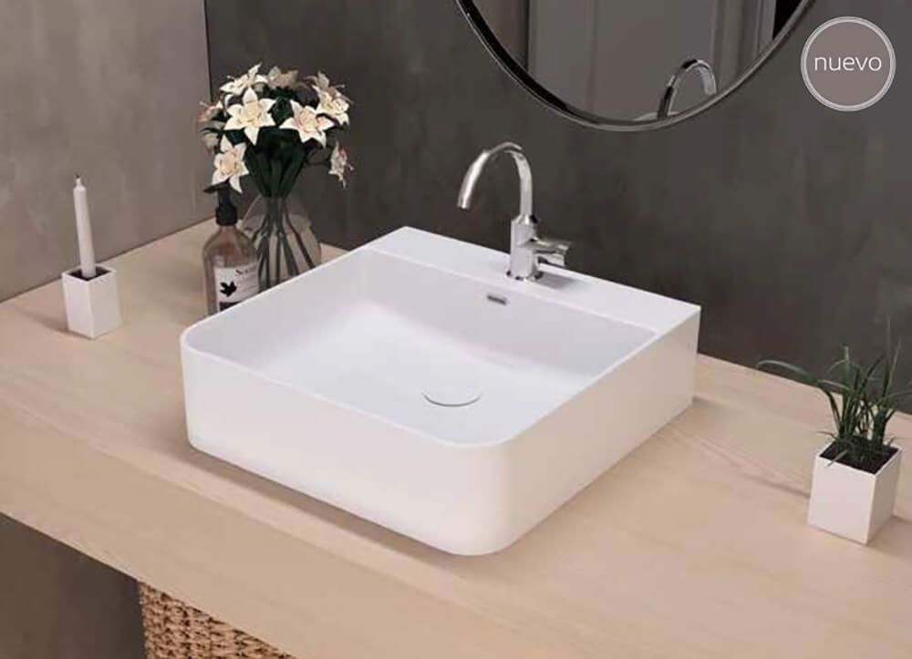 Lavabo de baño cuadrado blanco sobre encimera de madera clara con grifo cromado, flores y espejo.