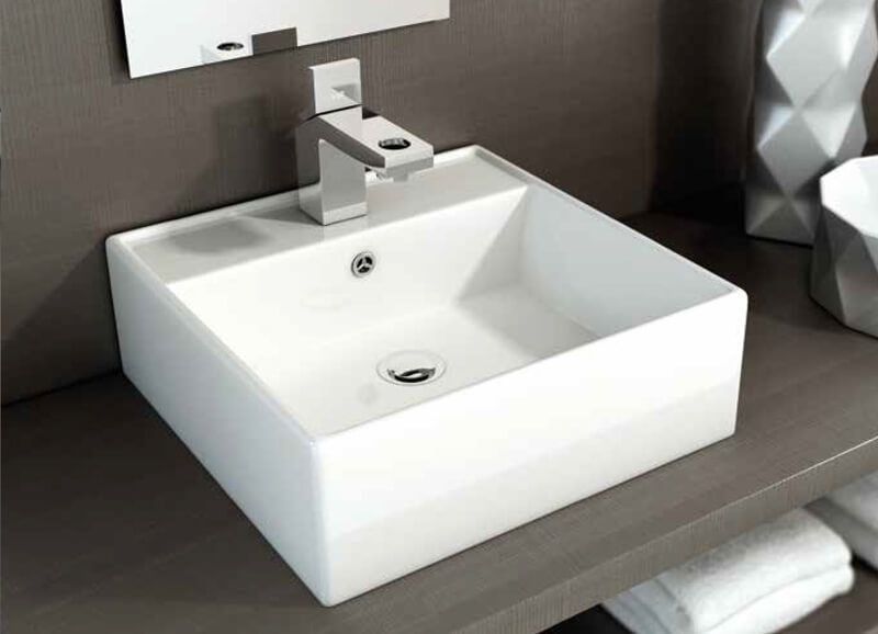 Lavabo cuadrado blanco con grifo cromado, colocado sobre un estante. Al fondo, una toalla blanca y una decoración.