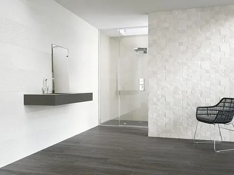 Baño moderno con tocador flotante, ducha de cristal y pared blanca texturizada. Suelo oscuro y silla negra.