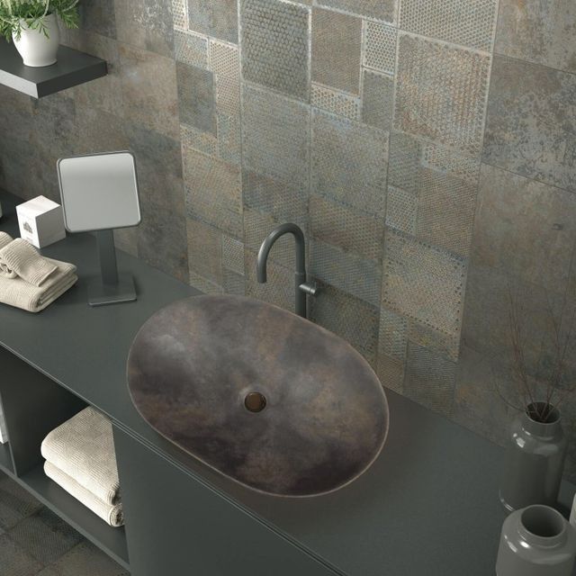 Baño con pared de baldosas de piedra, lavabo ovalado y encimera gris oscuro con estante.