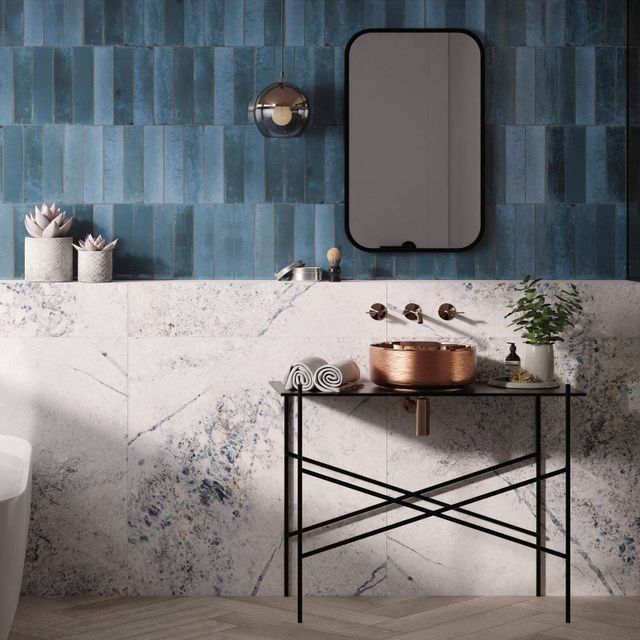 Baño con azulejos azules, lavabo de cobre, espejo negro y soporte negro.