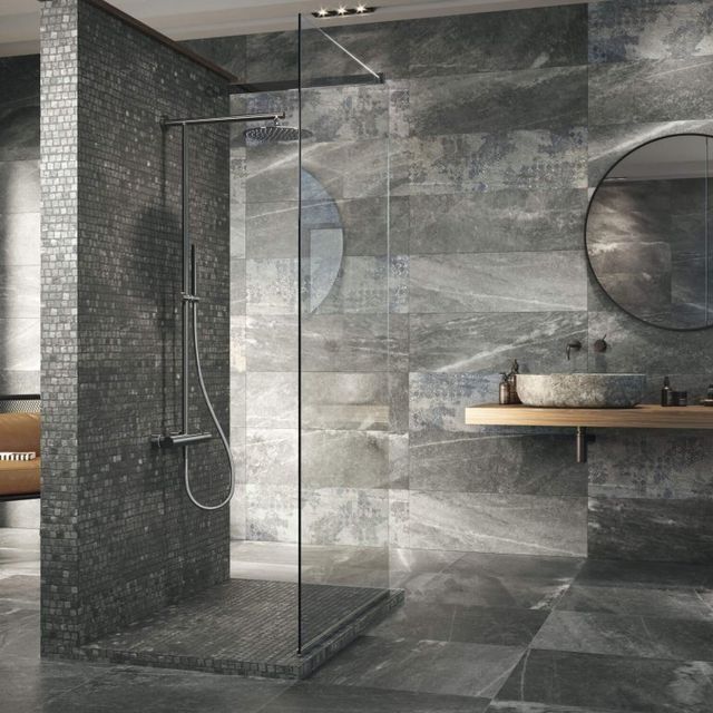 Baño moderno con paredes de azulejos grises, ducha y espejo circular.