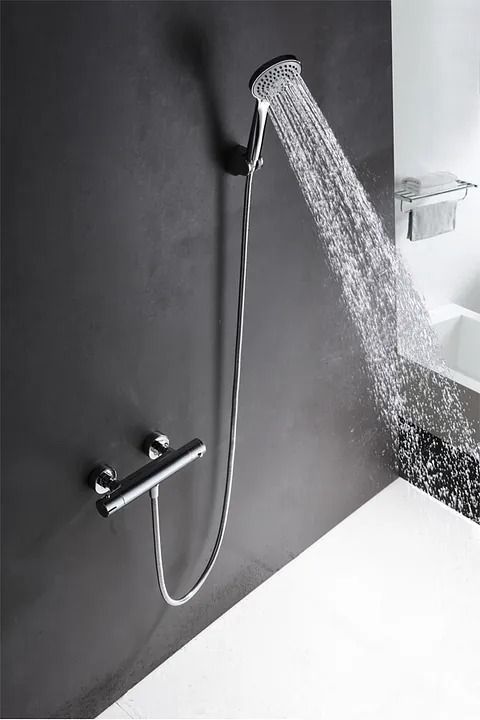 Cabezal de ducha que rocía agua contra una pared gris oscuro; lavabo blanco en el fondo.