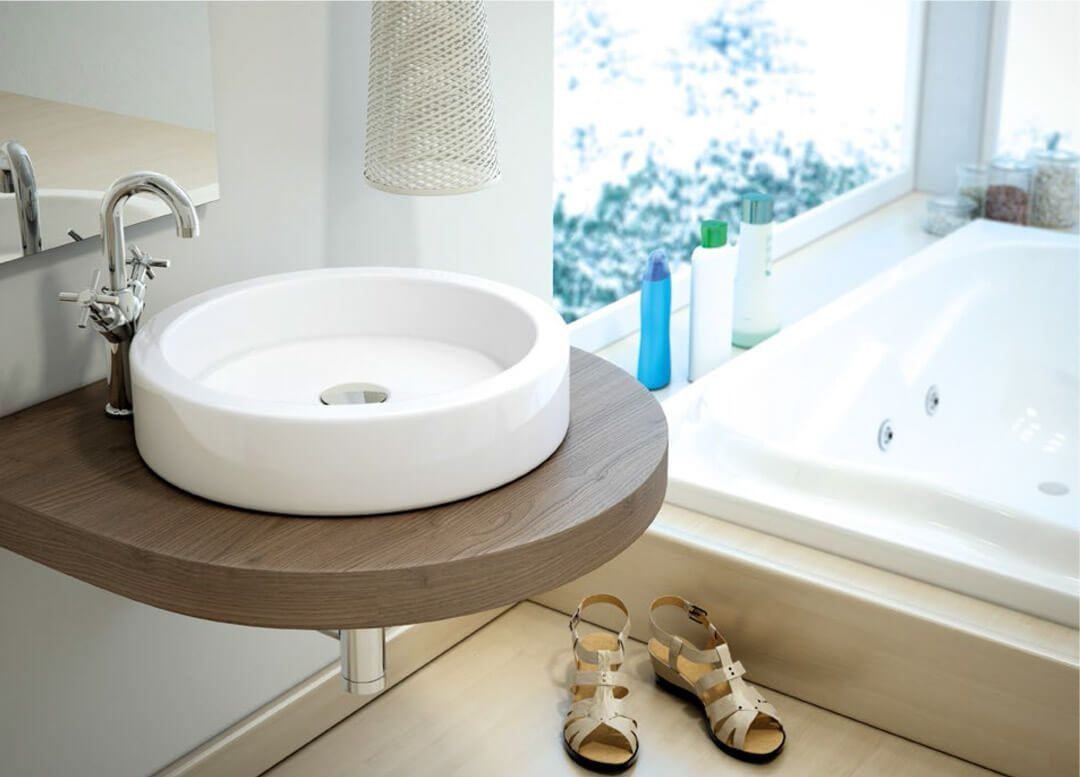Lavabo circular blanco sobre una repisa de madera en un baño con ventana y bañera. Sandalias beige en el suelo.