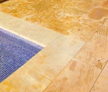 Terraza de piscina de piedra amarilla con una esquina de piscina con azulejos azules; superficie húmeda.
