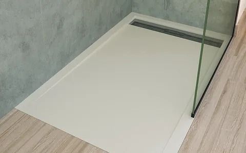 Plato de ducha blanco con desagüe lineal, mampara de cristal, suelo de madera clara y pared texturizada.