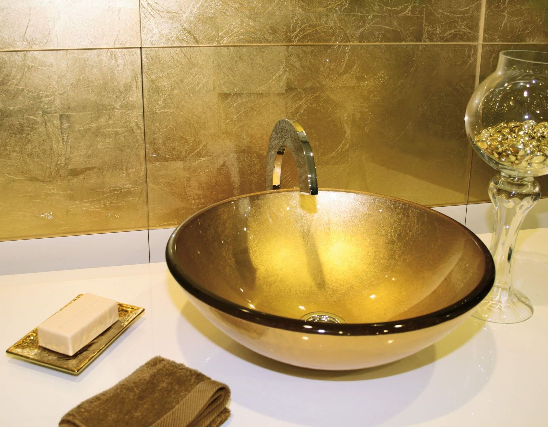 Lavabo de baño en tonos dorados con grifo y jabonera; fondo de azulejos con pan de oro.