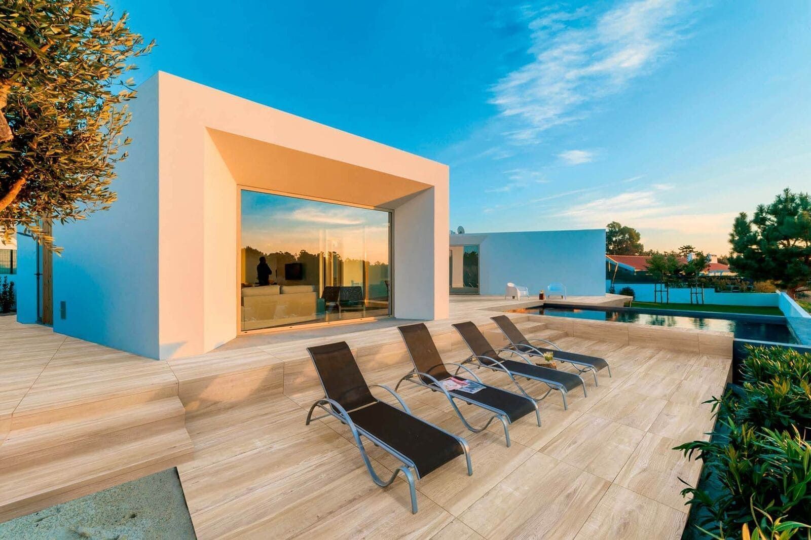 Edificio blanco moderno con puertas de cristal, tumbonas junto a una piscina en un patio de piedra, cielo azul.