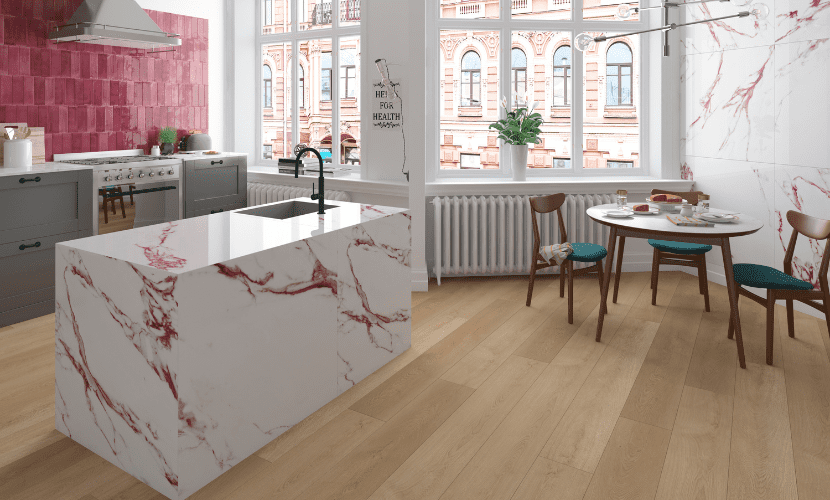 Cocina con isla de mármol blanco y vetas rojas, suelo de madera y salpicadero rosa. A la derecha hay una mesa redonda con sillas.