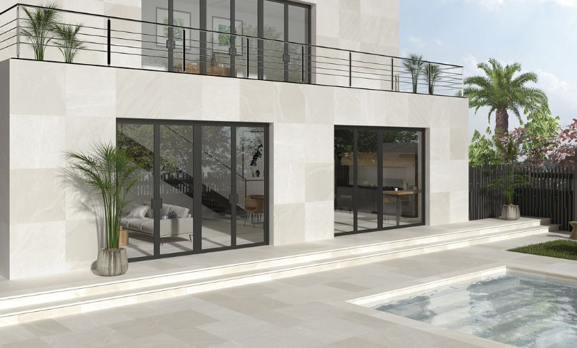 Casa moderna de dos plantas con fachada de piedra clara, puertas con marco negro, balcón y piscina. Palmeras y vegetación a su alrededor.