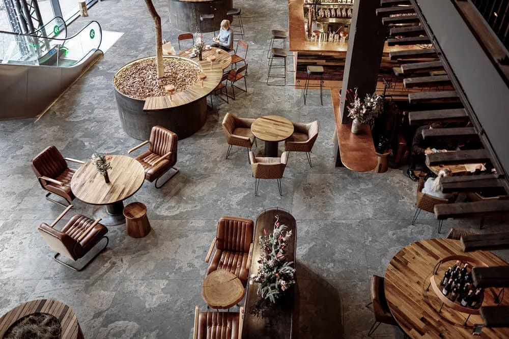 Vista aérea de un restaurante con mesas, sillas, barra y escalera mecánica. Diseño moderno con detalles en madera.