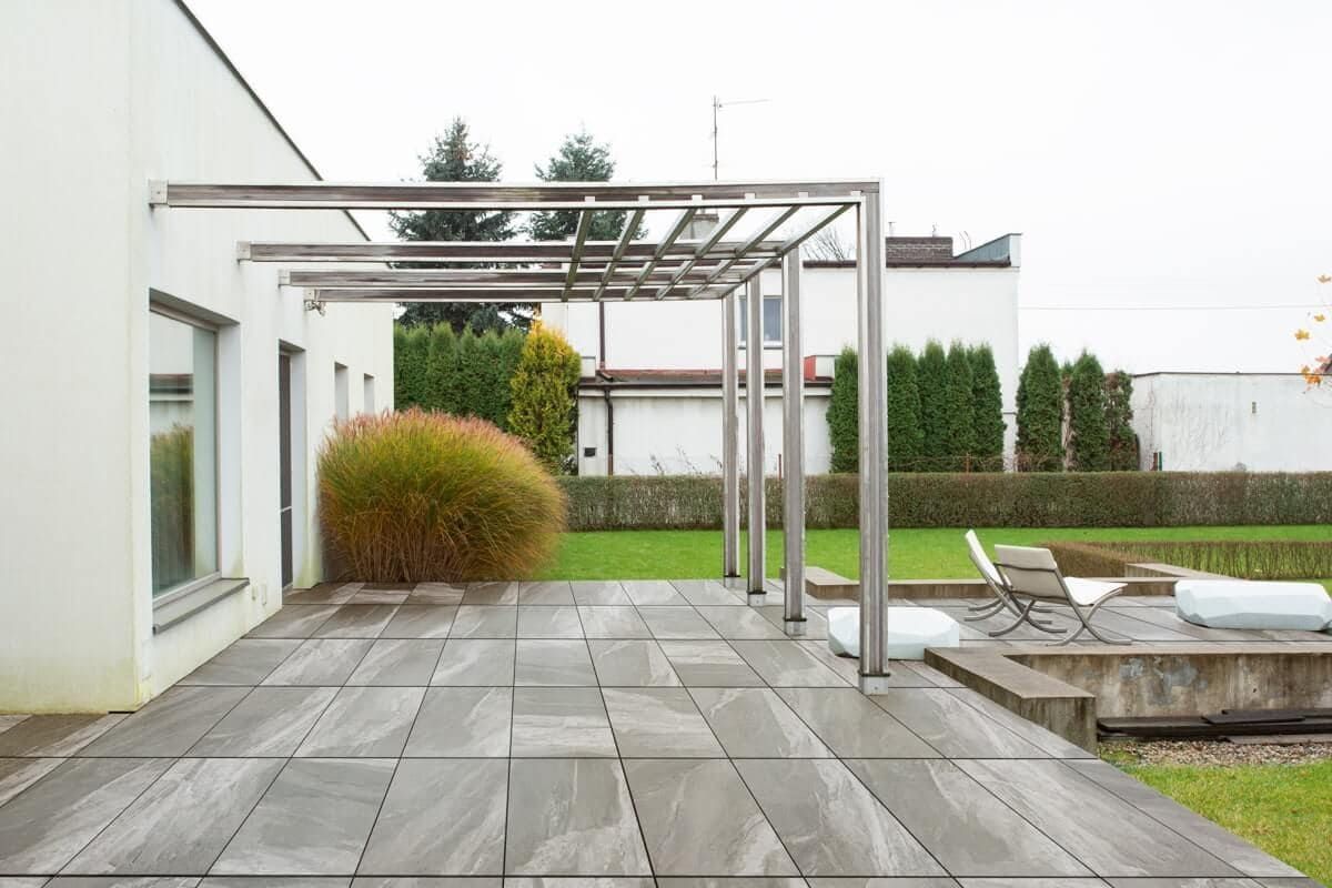Patio con baldosas grises, pérgola metálica, césped y edificio blanco.