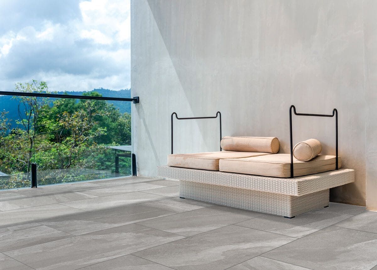 Patio al aire libre con diván tejido en color blanco, cojines beige y barandilla de cristal con vista a la montaña.