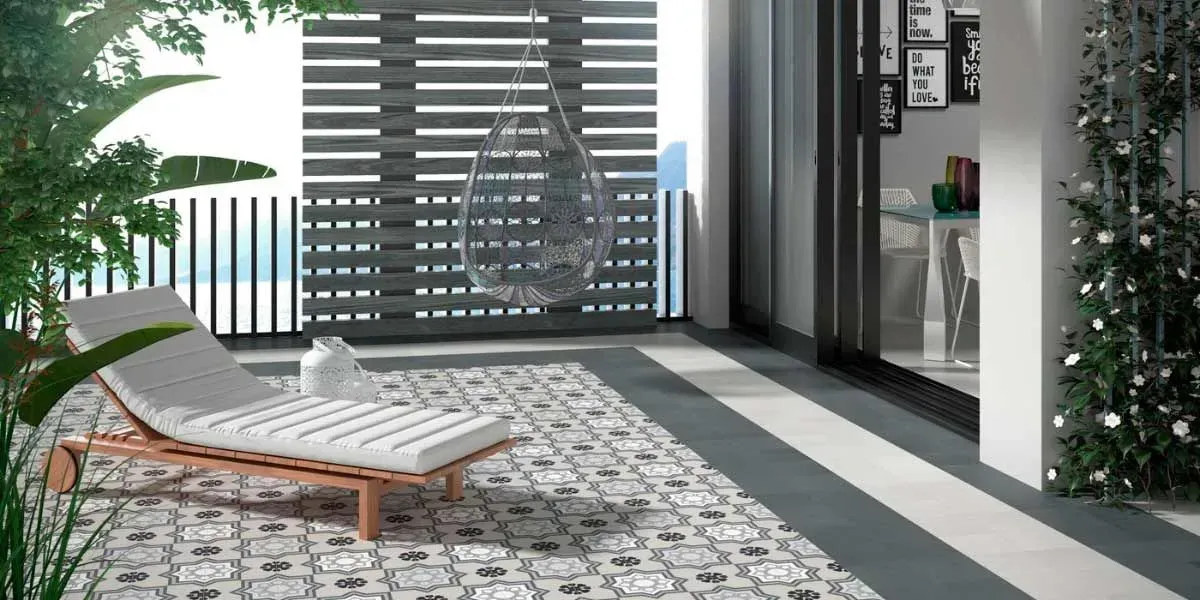 Patio al aire libre con un sillón blanco, azulejos estampados y una silla colgante, con vegetación.