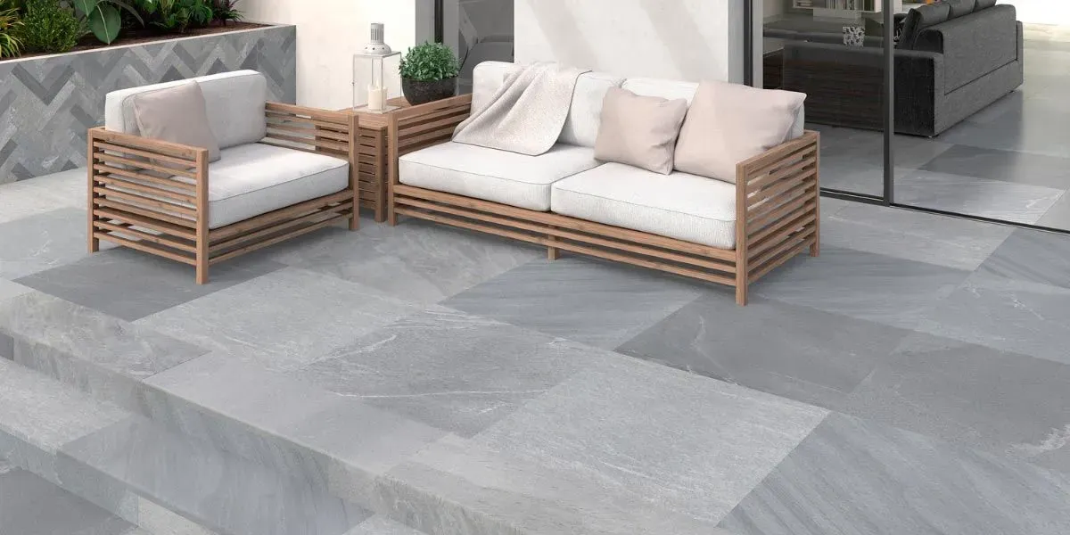 Muebles de patio sobre suelo de piedra gris. Dos sofás de madera con cojines blancos y una mesa auxiliar.