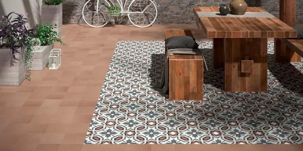 Patio exterior con mesa y bancos de madera sobre una alfombra estampada; plantas en macetas y una bicicleta al fondo.