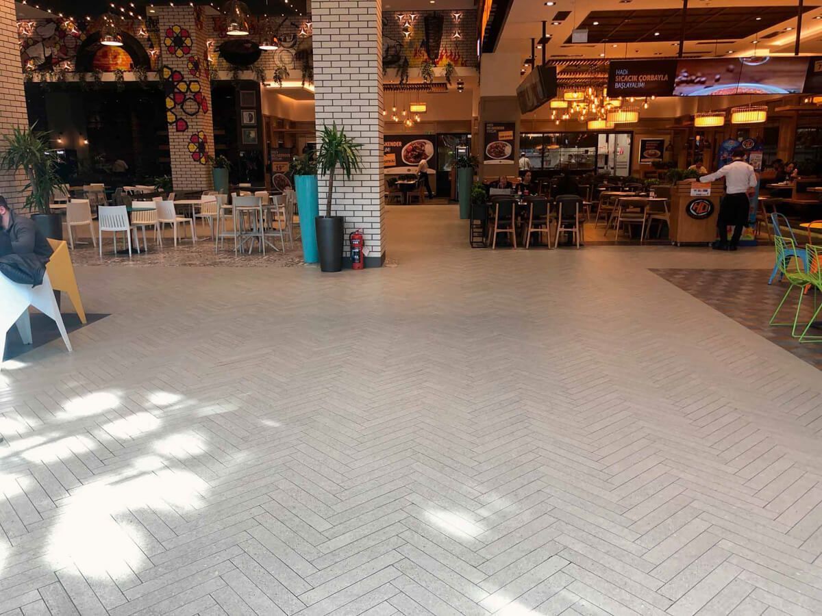 Amplio patio de comidas con piso de baldosas en espiga, mesas, sillas y múltiples puestos de restaurante.