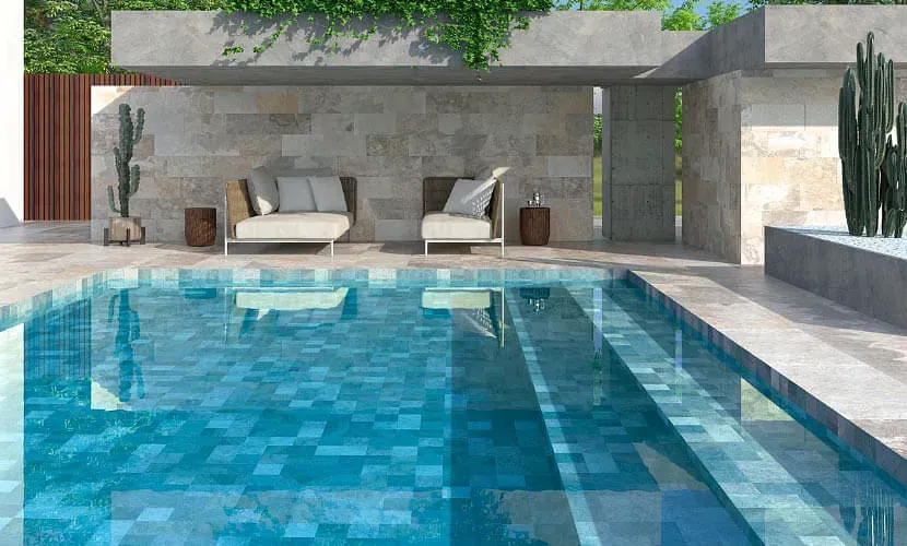 Piscina con agua de azulejos azules, escalones y tumbonas bajo un alero cubierto de piedras; cactus cerca.