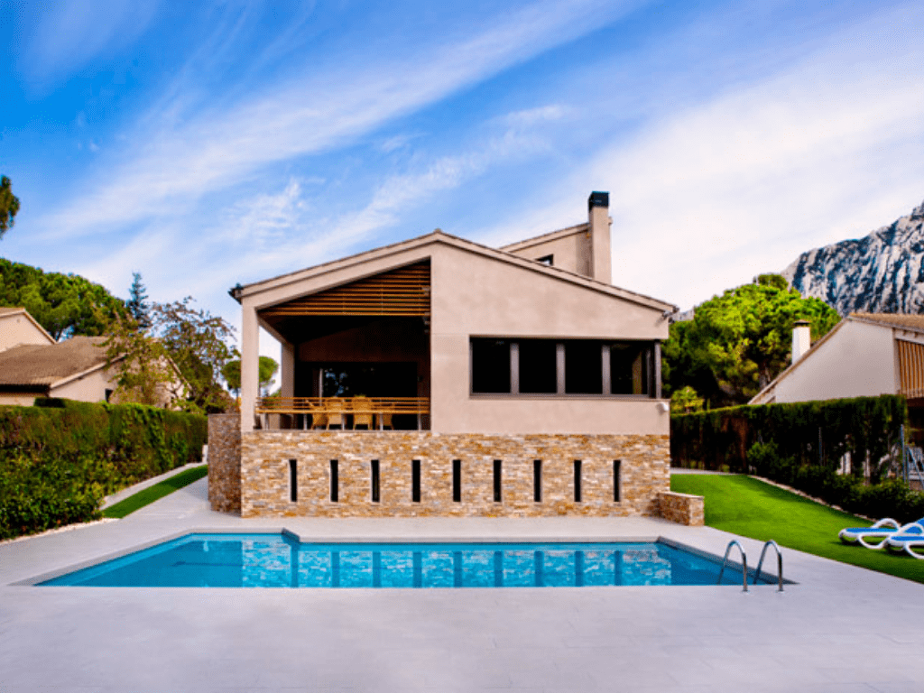 Casa moderna con piscina; fachada beige y piedra, deck de madera, piscina azul, césped verde, cielo azul.