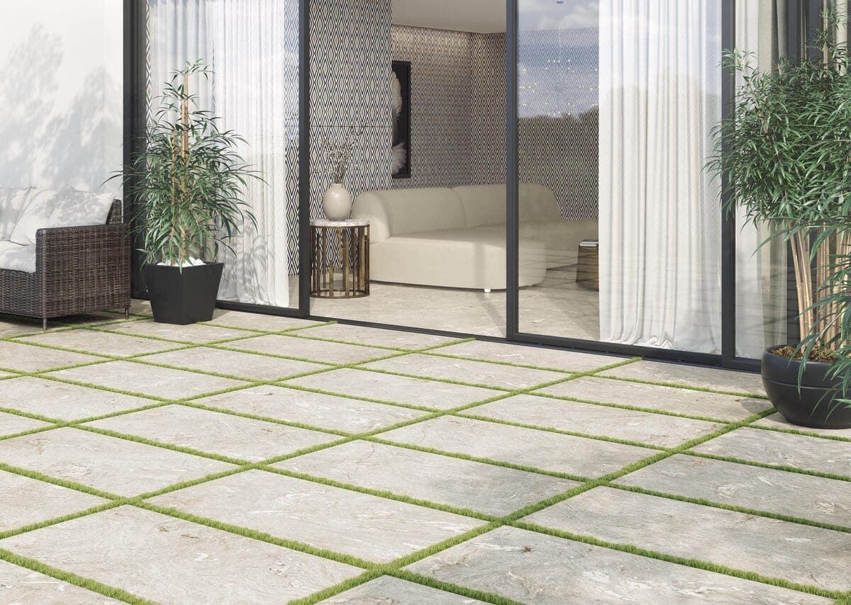 Patio con adoquines cuadrados y lechada verde que conduce a una habitación con puertas de vidrio y un sofá blanco.