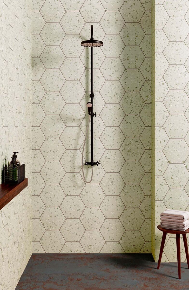 Ducha con grifería negra y azulejos con diseño hexagonal. Un estante de madera contiene botellas y se ve un taburete con toallas.