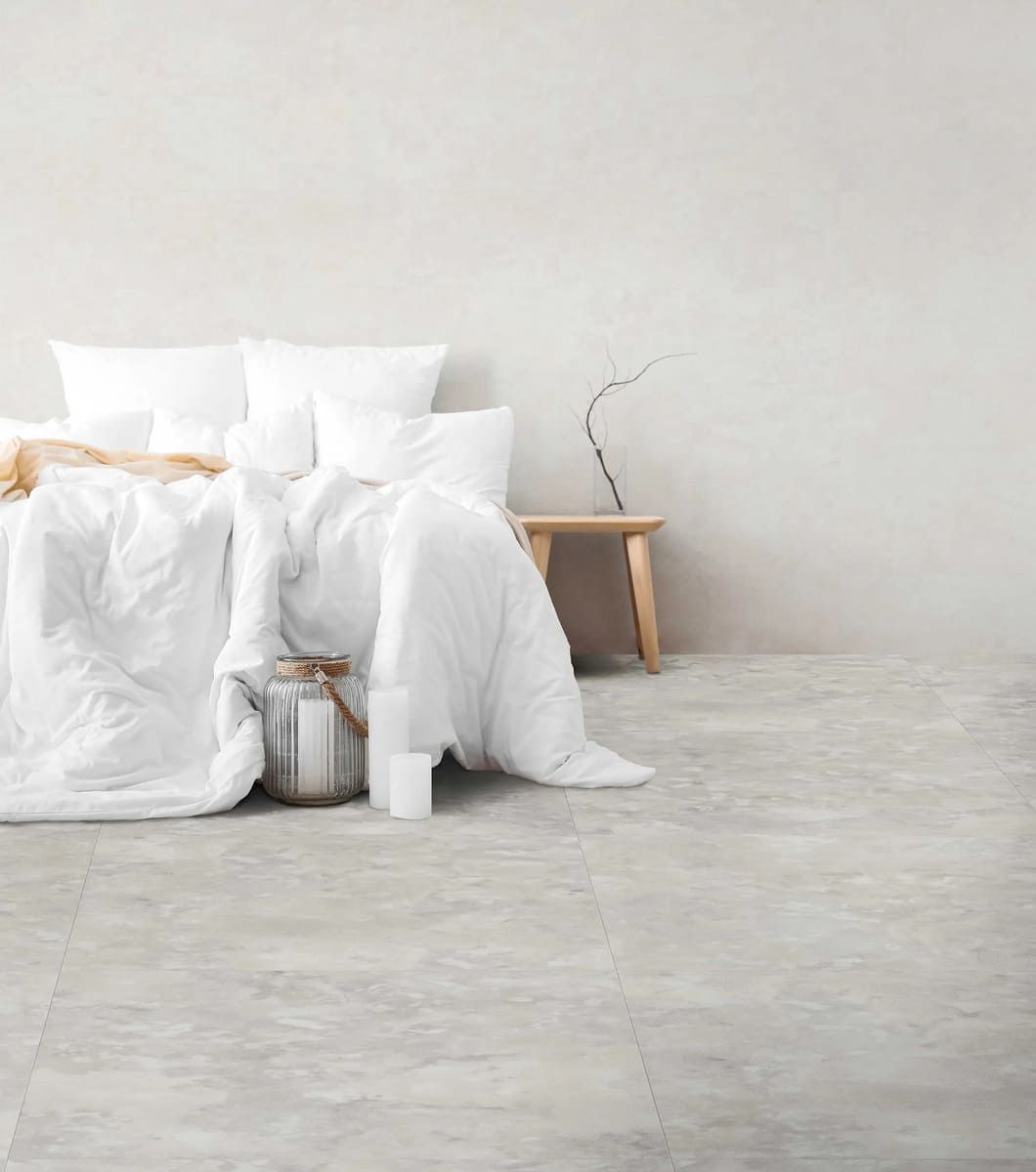 Dormitorio con cama, ropa de cama y paredes de color blanco, con taburete de madera y suelo gris.