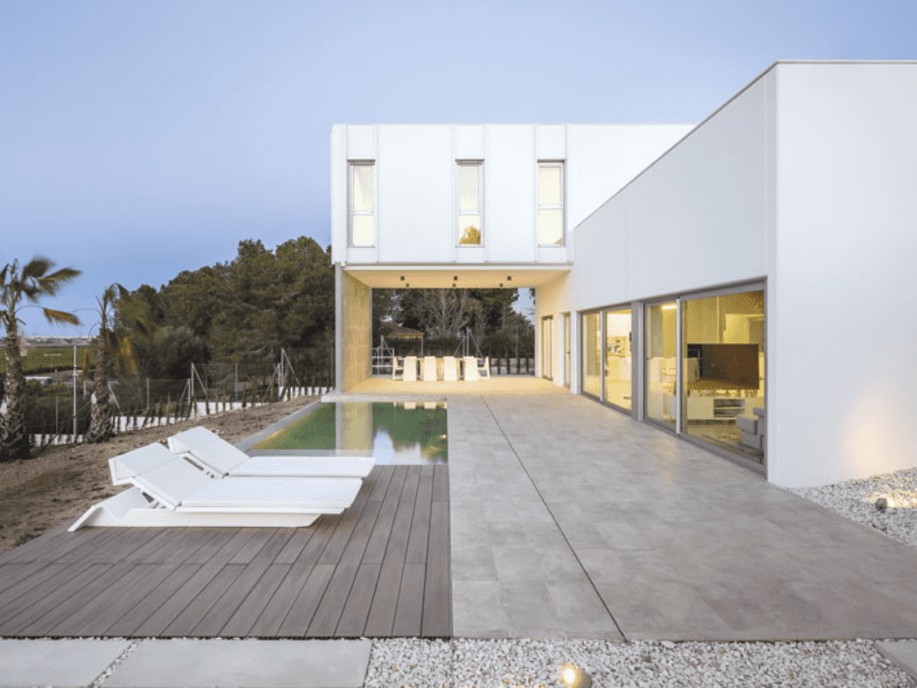 Moderna casa blanca con piscina, tumbonas y grandes ventanales.