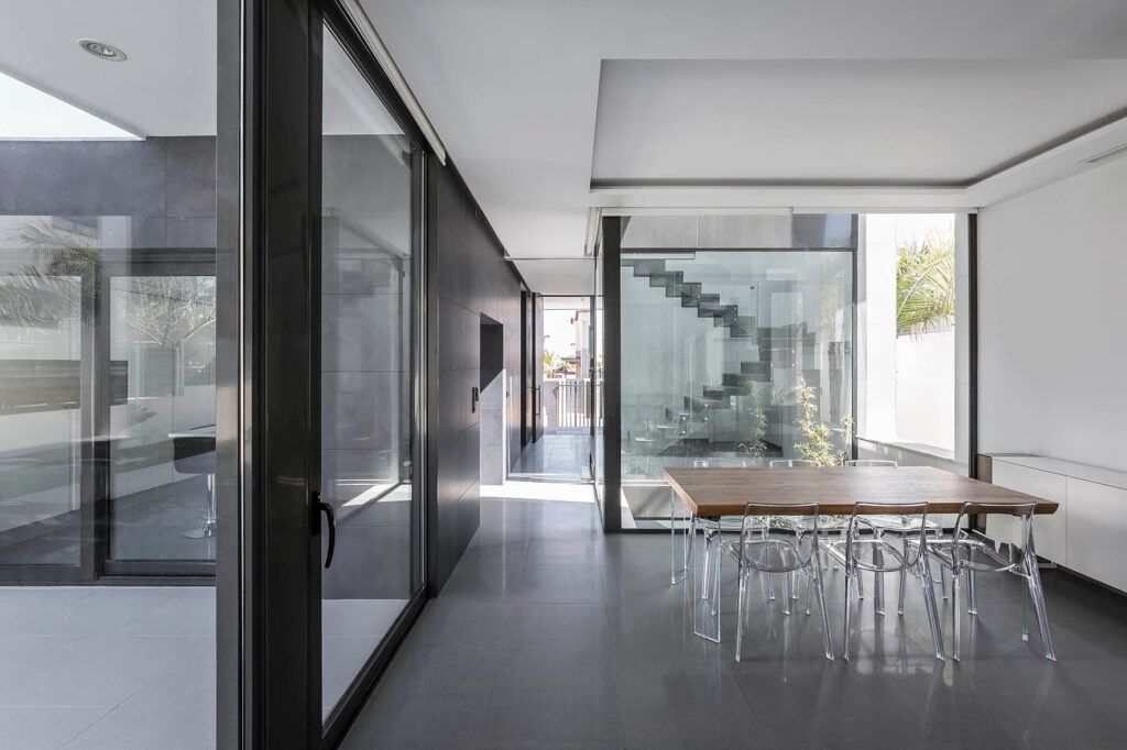 Interior moderno con puertas corredizas de vidrio, piso gris pulido, mesa de madera y sillas transparentes.