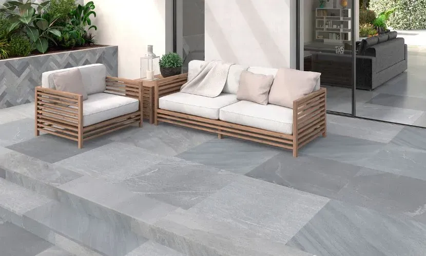 Patio con baldosas de piedra gris, asientos de madera con cojines blancos y vegetación.