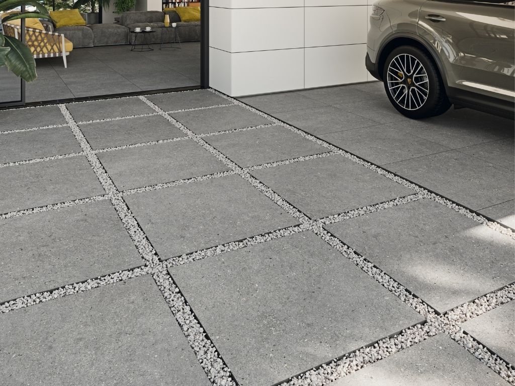 Pavimentos grises con grava en el medio, al lado de un coche y una entrada de casa moderna.