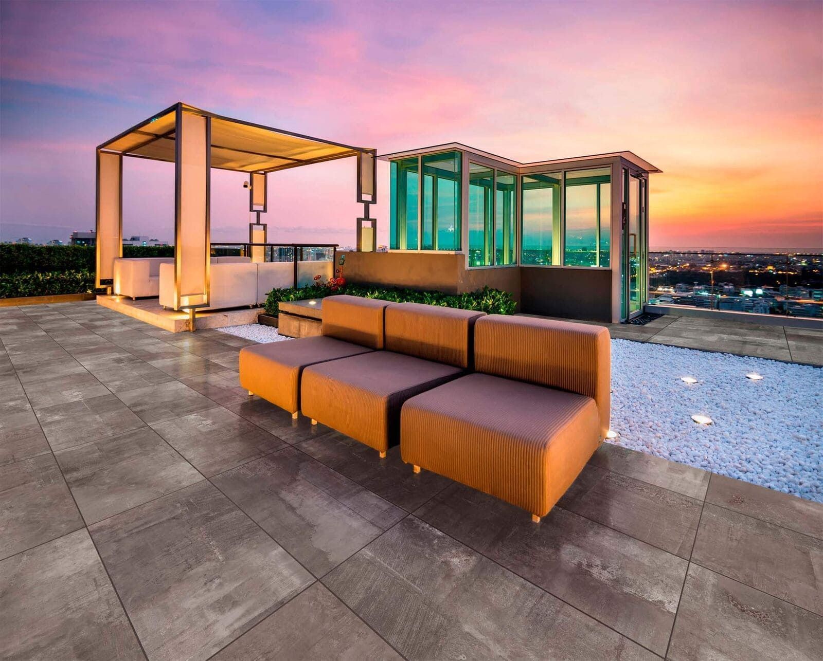 Salón en azotea al atardecer con sofá modular, marquesina, cerramiento de cristal y vista a la ciudad.