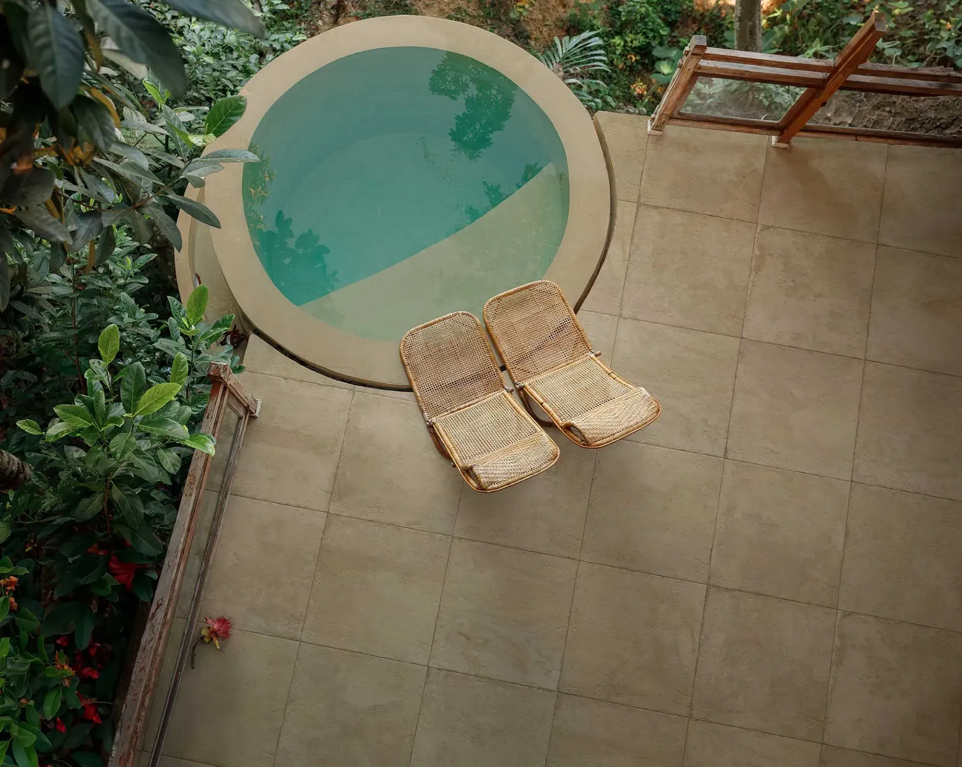 Piscina redonda con agua turquesa, dos sillas tejidas y terraza de concreto circundante en un exuberante entorno al aire libre.