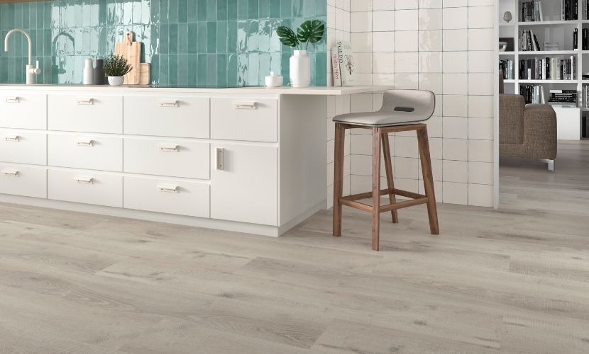 Cocina con gabinetes blancos, salpicadero verde azulado y suelo de madera clara. Hay un taburete junto a la encimera.