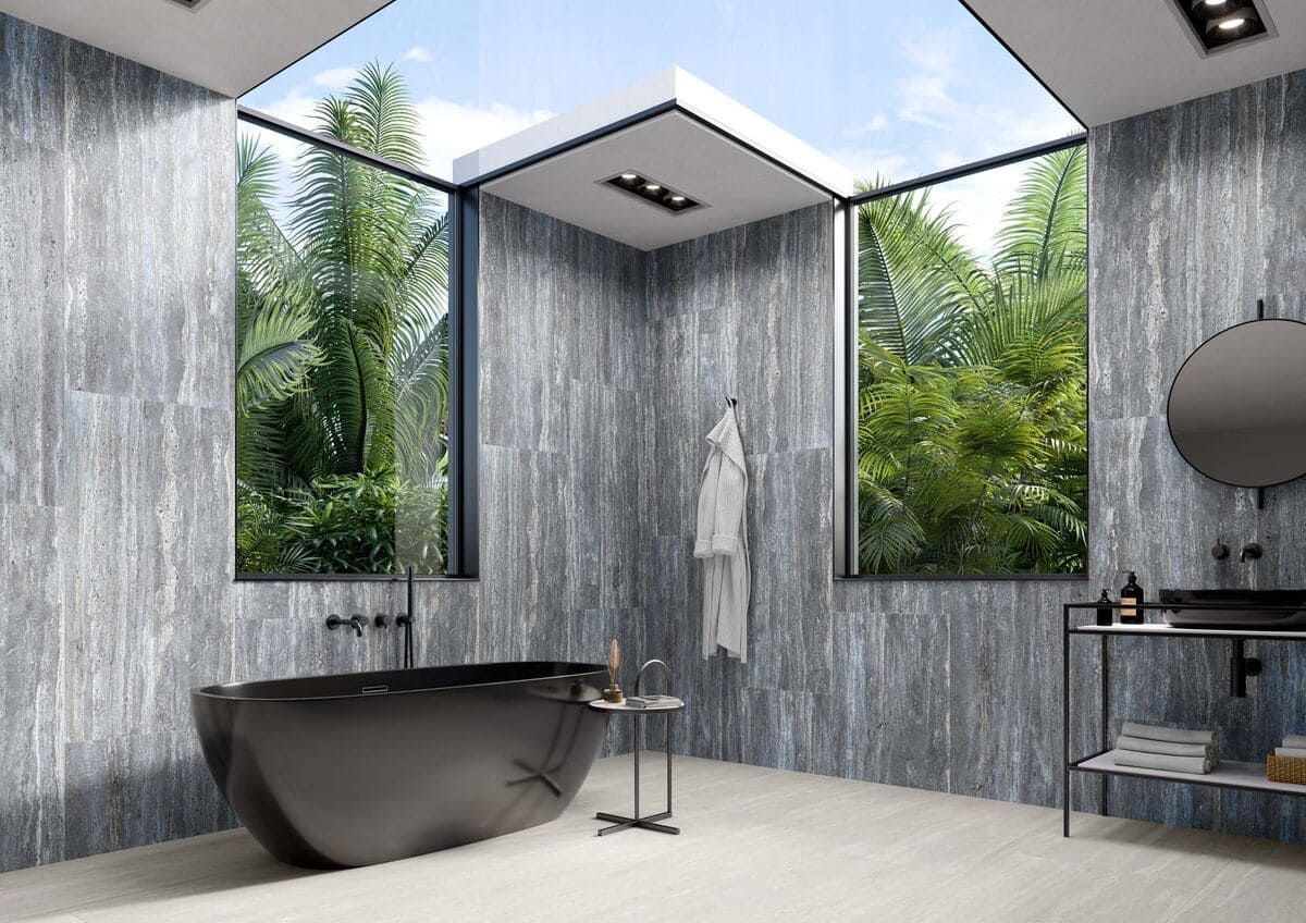 Baño moderno con bañera negra, grandes ventanales con vistas a la vegetación, paredes grises y tragaluz.