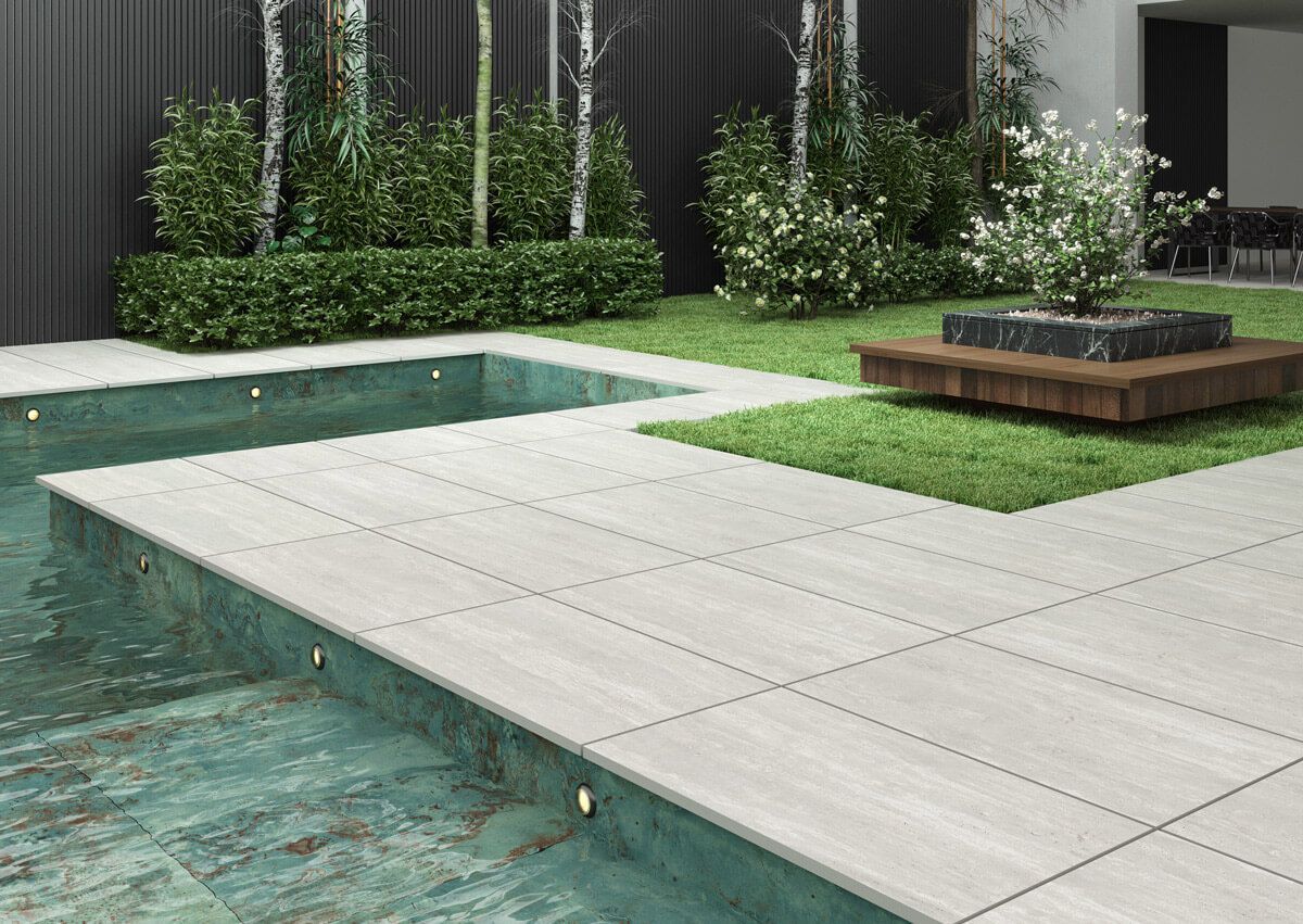 Patio junto a la piscina con baldosas de piedra gris claro, piscina turquesa, césped verde y vegetación.