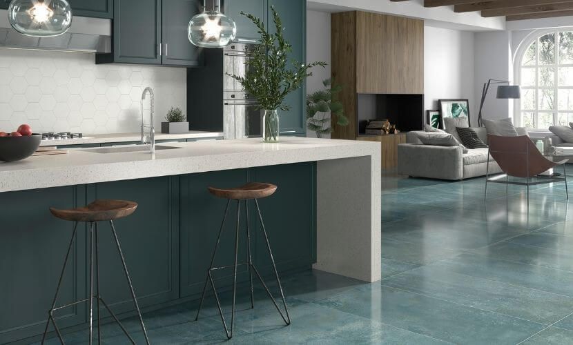 Cocina con gabinetes azules, encimeras blancas y taburetes de bar; sala de estar al fondo con chimenea y grandes ventanales.