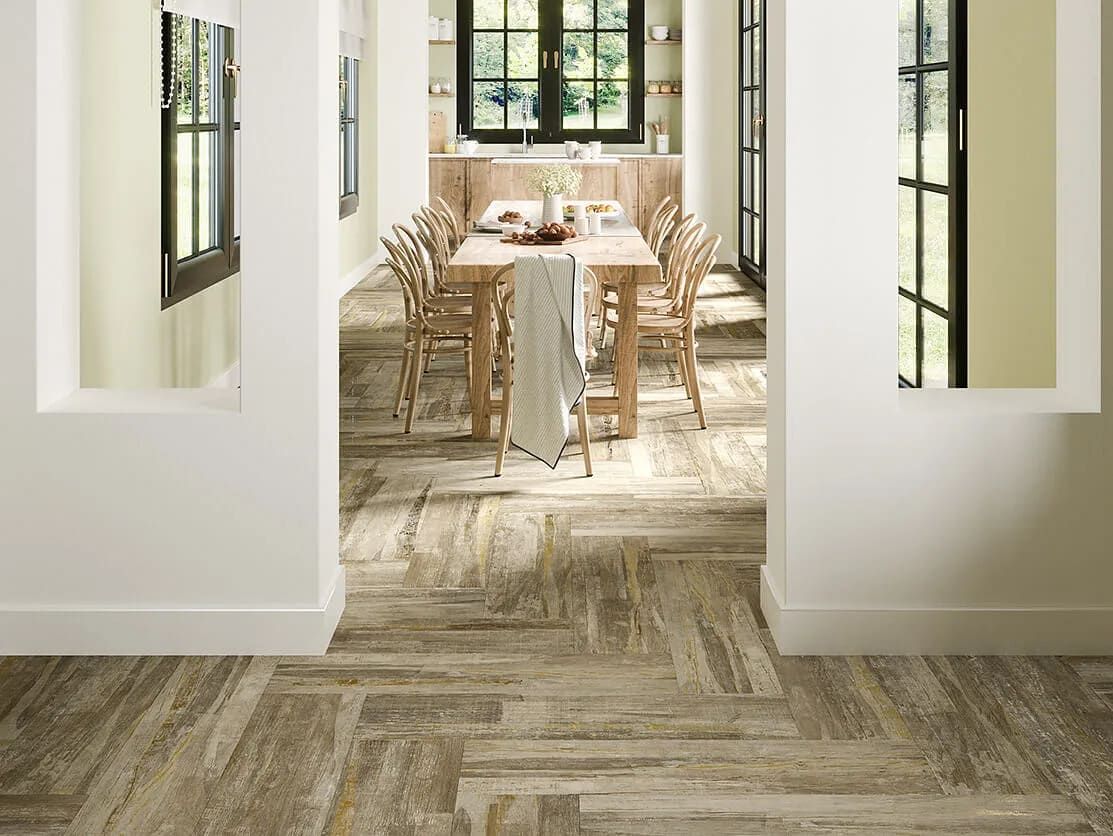 Comedor con una mesa larga de madera, sillas y suelo de espiga. Paredes y ventanas blancas.